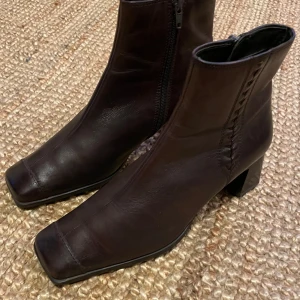 Bruna läder boots med fyrkantig tå - Snygga bruna boots i skinn med fyrkantig tå och markerad blockklack. Dragkedja på sidan för enkel påtagning och dekorativ söm längs skaftet. Köpta vintage, begränsat använda. Upplever dem som en stor/lång storlek 39, rymliga vid tårna pga fyrkantiga tån.