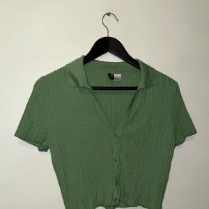 Grön ribbad croppad blus med krage - Snygg grön ribbad blus från H&M med korta ärmar, v-ringning och krage. Toppen är croppad och har knappar framtill, perfekt för en trendig look. Enkel att matcha med både jeans och kjol.