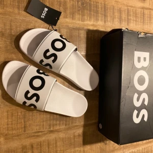 Vita sandaler från BOSS med logga - Snygga vita sandaler från BOSS med bred rem och stor svart BOSS-logga över foten. Tillverkade i syntetmaterial med platt sula och präglad logga på sidan. Perfekt för chill dagar och enkel att slänga på sig när du ska ut snabbt.