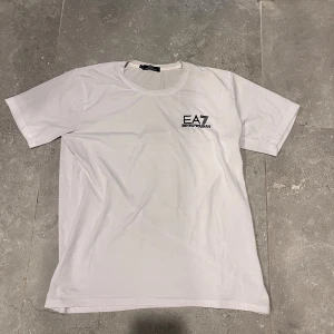 Vit EA7 t-shirt  - Snygg vit t-shirt från EA7 Emporio Armani med diskret svart logga på bröstet. Klassisk rund hals och korta ärmar, perfekt för en clean och stilren look. Mjuk bomull som känns skön mot huden. Passar dig som gillar exklusiva märken och enkel design.