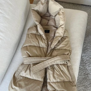 Beige Väst från Bershka - Superfin beige väst som passar perfekt nu till våren. Från märket Bershka. Storleken är Xs-S men den är oversize i modellen. Har luva , knappar & bälte i midjan. Aldrig använd som ny. Säljer för 200 kr.