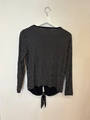 Prickig långärmad topp från Bodyflirt - Svart långärmad topp med vita prickar från Bodyflirt. Toppen har rund halsringning och en snygg knytning framtill som ger en cool detalj. Tillverkad i mjukt och stretchigt material som sitter skönt på kroppen.