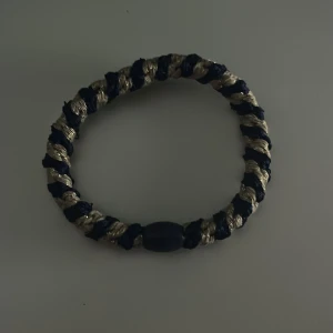 Blått och guldig knekki  - Snyggt flätat armband i mörkblått och guld med en oval pärldetalj. Armbandet har en grov, tvinnad design som ger en cool och unik look. Perfekt för dig som gillar att sticka ut med accessoarer.