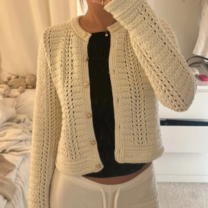 Beige stickad kofta med guldfärgade knappar - Superfin beige stickad kofta med snygga guldfärgade knappar framtill. Koftan har ett öppet hålmönster och rund hals, samt långa ärmar. Perfekt att slänga över en topp för en chill och stilren look.