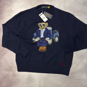Mörkblå stickad tröja Polo Ralph Lauren - Snygg mörkblå stickad tröja från Polo Ralph Lauren med ikonisk björn broderad på framsidan. Tröjan har ribbade muddar och rund hals. Tillverkad i mjuk bomull med RL-broderi i rött vid nederkanten. Perfekt för dig som gillar klassisk stil med en twist.