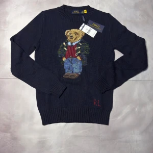 Mörkblå stickad tröja Polo Ralph Lauren - Mörkblå stickad tröja från Polo Ralph Lauren med ikonisk björn broderad på bröstet. Rund hals, ribbade muddar och RL-broderi nedtill. Materialmix av bomull och linne, perfekt för dig som gillar klassisk stil med en twist.
