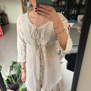 Beige spetsklänning från Culture - Supersöt kortklänning i beige från Culture med broderade blommor, spetsdetaljer och knappar framtill. Klänningen har trekvartsärm, snörning vid halsen och en luftig passform. Perfekt för sommarens alla tillfällen!