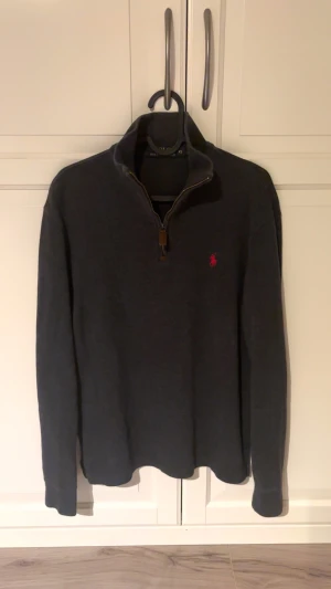 Mörkgrå half zip tröja från Polo Ralph Lauren - Snygg mörkgrå långärmad tröja från Polo Ralph Lauren med röd broderad logga på bröstet. Tröjan har en half zip-dragkedja med läderdetalj och hög krage. Perfekt för dig som gillar klassisk stil med en modern touch. 9/10