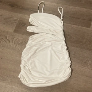 Vit omlott kortklänning med cut-out - Säljer en vit kortklänning med smala axelband och snygg cut-out detalj i midjan. Klänningen har omlott-design och är figurnära med lätt rynkade sidor. Perfekt för dig som gillar trendiga och stilrena plagg med lite edge.