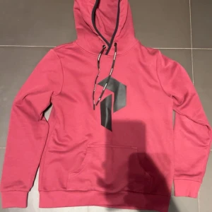 Röd hoodie från Peak Performance - Snygg röd hoodie från Peak Performance med svart logga på bröstet och svarta snören med märkets namn. Tröjan har huva, magficka och ribbade muddar. Perfekt för chill eller häng med kompisar. Materialet är mjuk bomull och polyesterblandning.