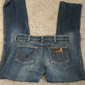 Wrangler blå bootcut jeans - Snygga blå jeans från Wrangler med klassisk bootcut passform. Jeansen har kontrastsömmar, fem fickor och detaljerad brodering på bakfickorna. Märkespatch i mocka på bakfickan och metallknapp med Wrangler-logga. Perfekt för dig som gillar tidlös denimstil.