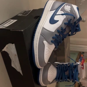 Nike Air Jordan 1 Mid blå/grå - Nike Air Jordan 1 Mid sneakers i en clean färgway med vit bas, gråa paneler och blå detaljer. Swoosh och snörning i blått, samt blå yttersula. Klassisk Jordan Wings-logga på sidan. Skorna har perforerad tå och är tillverkade i skinn för en snygg och sportig look.