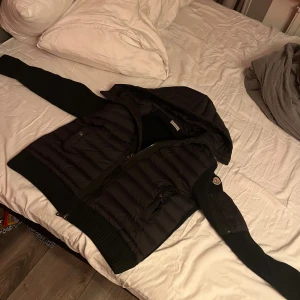 Svart Moncler pufferjacka med huva - Svart pufferjacka från Moncler med stickade ärmar och rygg, quiltad framsida och huva. Jackan har dragkedja framtill, fickor med tryckknapp och en Moncler-logga på ena ärmen. Perfekt för kyliga dagar och riktigt snygg streetstil.