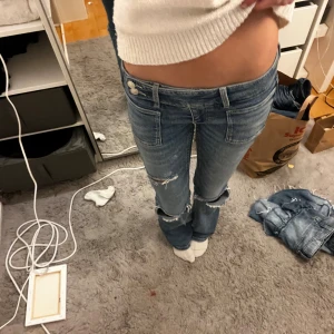 Blå bootcut jeans med slitningar - Snygga blå jeans med bootcut passform och låg midja. Jeansen har slitna detaljer och hål vid knäna för en cool, avslappnad vibe. Framtill finns stora fickor och klassiska bakfickor. Perfekta för dig som gillar en trendig och edgy stil.