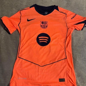 Barça Pedri 8 Nike fotbollströja orange - Säljer en officiell FC Barcelona fotbollströja från Nike i orange med mörkblå detaljer. Tröjan har korta ärmar, klubbmärke, sponsorlogga (Spotify) och namn samt nummer (Pedri 8) på ryggen. Tillverkad i lätt och ventilerande material, perfekt för match eller träning.