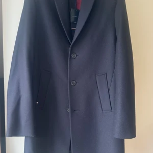 Mörk blå rock från Tommy Hilfiger, M - Snygg mörk blå  rock från Tommy Hilfiger i storlek M. Klassisk design med tre knappar framtill, två snedställda fickor och röd insida. Tillverkad i ullblandning som ger en lyxig känsla. Storlek 48-50, M                                                Stilren, 75% ull                                                        Helt ny, oanvänd 