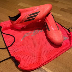 Adidas F50 rosa fotbollsskor - Säljer ett par Adidas F50 fotbollsskor i en riktigt snygg och neonrosa färg med svarta detaljer och klassiska tre ränder på sidan. Skorna har platt sula med dobbar för gräsplan och är tillverkade i lätt syntetmaterial. Medföljer matchande rosa F50-påse. Köptes för 3000 kr använda cirka 5 gånger på träning. Priset kan sänkas vid snabb affär