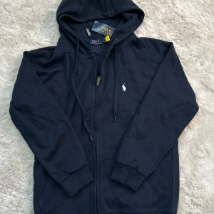 Blå hoodie från Polo Ralph Lauren - Snygg marinblå hoodie från Polo Ralph Lauren med klassisk broderad logga på bröstet. Tröjan har huva med snörning, dragkedja framtill och ribbade muddar. Perfekt för dig som gillar stilrena och bekväma plagg med premiumkänsla. Använd fåtal gågånger!! Priset kan diskuteras!