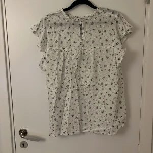 Vit blommig kortklänning Vero Moda - Supersöt vit blus från Vero Moda med små blå och gröna blommor. Klänningen har volangdetaljer vid ärmarna, spets och smock i ryggen samt rund halsringning. Tillverkad i lätt och skön bomull, perfekt för varma dagar. Säljes i storlek L.