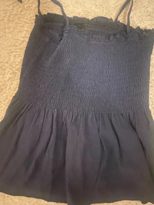 Svart smockad linnetopp med knytband - Svart linnetopp med smockad överdel och volangig nederdel. Toppen har tunna axelband som knyts upptill för en söt och justerbar passform. Perfekt till varma dagar och enkel att styla med jeans eller kjol.