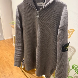 Grå stickad kofta från Stone Island - Säljer en grå stickad kofta från Stone Island med dragkedja och hög krage. Tröjan har långärmad passform och den klassiska Stone Island-loggan på vänster ärm. Ribbad struktur och snygg detalj på dragkedjan.