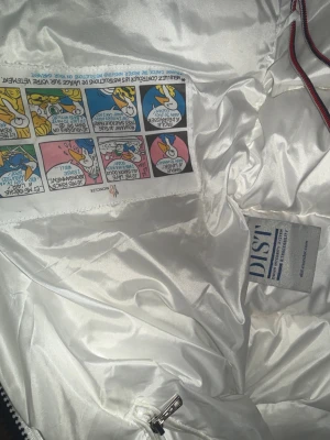 Vit dunväst från Moncler med huva - Säljer en snygg vit dunväst från Moncler med huva och dragkedja. Västen har Moncler-logga på bröstet, broderad text på huvan och detaljer med rödblå rand längs kanten. Fylld med dun och har praktiska fickor samt snörning vid huvan. Perfekt för kyliga dagar. Västen har inga defekter alls.