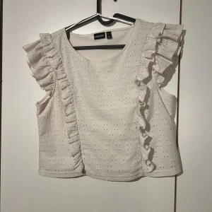 Vit blus med volang från Pieces - Supersöt vit blus från Pieces med broderade detaljer och volanger längs axlarna. Ärmlös modell med rund halsringning och lätt struktur i tyget. Perfekt för dig som gillar romantisk och feminin stil.