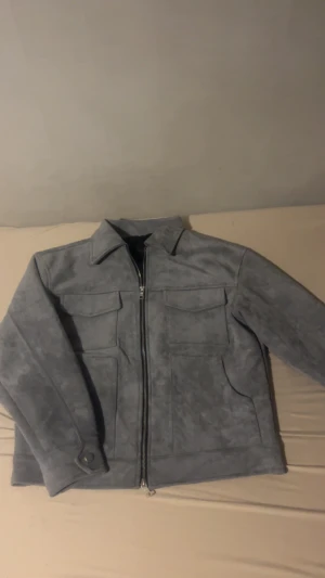 Grå mockajacka med dragkedja - Snygg grå suede jacka med klassisk krage och raka linjer. Jackan har två stora bröstfickor med lock, sidofickor och en hel dragkedja framtill. Materialet är mjukt och matt, vilket ger en cool vibe. Passformen är normal och jackan har långa ärmar med knappdetalj vid mudden.