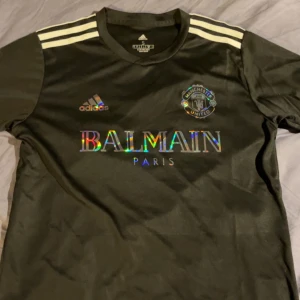 Manchester United x Balmain fotbollströja - Snygg svart Manchester United fotbollströja i samarbete med Balmain Paris. Tröjan har Adidas-logga, klubbmärke och holografiskt Balmain-tryck på bröstet samt vita ränder på axlarna. Tillverkad i ett lätt och sportigt material.