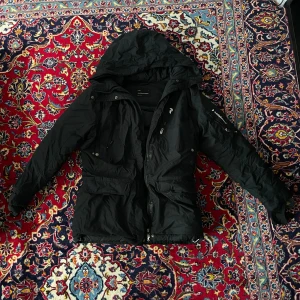 Svart Peak Performance Vinterjacka / Parkajacka | Storlek Large (UNISEX) | SWD Parka - Denna svarta, varma vinterjacka från PP är riktigt tjock, har handskydd, flera fickor och är designad för kalla dagar. Jackan är perfekt för svensk vinter.  ENDA PROBLEMET: Huvuddragkedjan är trasig. Du kan fortfarande stänga jackan lätt med dem svarta klisterlapparna (eller så kan man laga den och sätta in en ny dragkedja) - därför får du den väldigt billigt. Den har annars inga andra problem och har legat oanvänd länge.  Jackan är Unisex (både tjejer och killar kan bära den bekvämt).