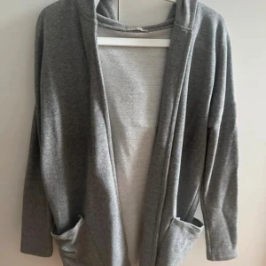 Grå hoodie-kofta med huva och fickor - Mysig grå hoodie-kofta med stor huva och två praktiska fickor framtill. Långärmad och öppen framtill, perfekt för lager-på-lager och chill dagar. Enkel och clean design som funkar till många stilar