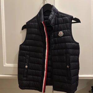 Svart dunväst från Moncler - Snygg svart dunväst från Moncler med quiltad design och ståkrage. Västen har två fickor med tryckknappar, dragkedja framtill och Moncler-logga på bröstet. Insidan har en vit etikett med Moncler-tryck. Röd, vit och blå rand längs dragkedjan ger en sportig touch. Storlek är 12 år (152 cm)