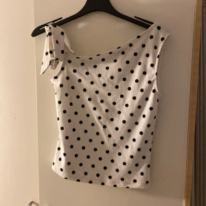 Prickig one shoulder topp från new yorker  - Snygg vit topp med svarta prickar från new yorker. One shoulder-modell med knyt vid axeln och figurnära passform. Perfekt för dig som vill sticka ut med en trendig och lekfull look.