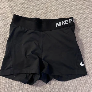 Nike Pro svarta träningstights shorts - Svarta Nike Pro shorts i tight modell med elastisk midja och vit Nike-logga på benet. Perfekta för träning eller sport, tillverkade i stretchigt syntetmaterial som andas. Snygg och sportig look med diskret logga på linningen.