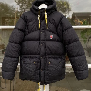 Svart pufferjacka från Fjällräven XS - Svart pufferjacka från Fjällräven med gul snörning i huvan, broderad logga på bröstet och stora fickor med knappar. Jackan har dragkedja och tryckknappar framtill, samt en skön huva för extra värme. Perfekt för kalla dagar.