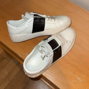 Valentino Open sneakers vit/svart - Snygga Valentino Open sneakers i vitt skinn med bred svart panel över sidan och klassiska nitar längs sulan. Modellen har rund tå, platt sula och snörning framtill. Perfekt för dig som gillar stilrena och lyxiga sneakers med en edgy touch.