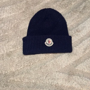 Mörkblå Moncler Mössa - Snygg mörkblå mössa från Moncler med klassisk uppvikt kant och broderad logotyp framtill. Perfekt för dig som vill ha en stilren och exklusiv look under kyliga dagar. Materialet är mjukt och värmande ull.