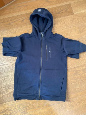 Marinblå hoodie från Sail Racing - Snygg marinblå hoodie från Sail Racing med hel dragkedja och huva. Storlek 170 cm. Tröjan har en bröstficka med dragkedja och logga tryckt på framsidan. Mjuk bomullskvalitet, perfekt för chill dagar. Klassisk passform och stilren design.