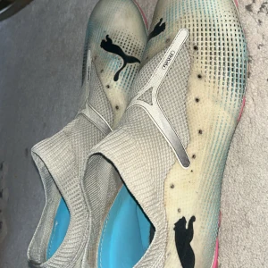 Puma fotbollsskor med blå detaljer - Säljer ett par gråa och ljusblå fotbollsskor från Puma med stickad ovandel och svart Puma-logga. Skorna har en strumpliknande passform, rosa dobbar och grafiska detaljer på sidan. Perfekt för dig som vill ha snygga och bekväma skor på planen.