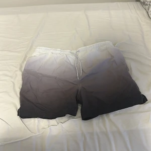 Svart-vita badshorts från Primark - Säljer ett par snygga badshorts från Primark i storlek M. De har en grym gradient från vitt upptill till svart nertill, elastisk midja med snörning och meshfoder. Perfekta för stranden eller poolen!