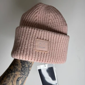 Ljusrosa ribbad mössa från Acne Studios i 100% ull - Snygg ljusrosa mössa från Acne Studios i 100% ull. Ribbstickad design med bred uppvikt kant och Acne Studios klassiska ansiktslogga framtill. Perfekt för dig som vill ha en clean och trendig look under kyliga dagar.