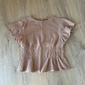 Beige blus med volang Xs - Superfin beige blus från SHEIN i storlek XS. Toppen har korta ärmar, markerad midja med smock och volang nertill. Tillverkad i ett mjukt, strukturerat bomullstyg som känns luftigt och skönt. Perfekt till jeans eller kjol för en chill look.