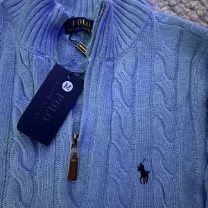 Blå kabelstickad tröja Polo Ralph Lauren - Kabelstickad blå tröja från Polo Ralph Lauren med halv dragkedja och ribbad krage. Klassisk broderad logga på bröstet och snygga detaljer vid dragkedjan. Perfekt för dig som gillar preppy stil och vill ha något både bekvämt och stilrent.