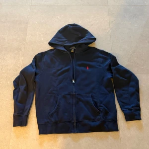 Marinblå hoodie från Polo Ralph Lauren - Snygg marinblå hoodie från Polo Ralph Lauren med röd logobrodyr på bröstet. Tröjan har huva, dragkedja framtill och två stora fickor. Tillverkad i mjuk bomullsmix som är perfekt för chill dagar. Klassisk och stilren design som funkar året runt. Några frågor är de bara och fråga 👍