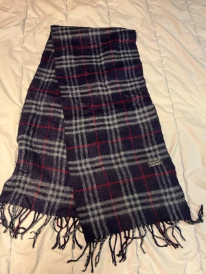 Rutig ullhalsduk från Burberrys - Mörkblå och grå rutig halsduk från Burberrys med röda detaljer. Gjord i 100% lammull och har klassiska fransar i ändarna. Perfekt accessoar för att lyfta din outfit med en tidlös brittisk känsla. N35