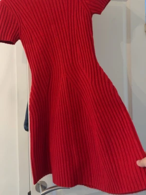 Röd ribbad stickad kortklänning - Superfin röd stickad klänning med ribbat mönster och korta ärmar. Klänningen har en figurnära passform upptill och svänger ut nertill för en snygg siluett. Perfekt för dig som vill ha något bekvämt men ändå stilrent. Helt ny!!