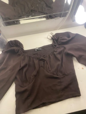 Brun blus med puffärmar från Bik Bok - Säljer en brun blus från Bik Bok i storlek M. Toppen har puffiga långärmar, smockad rygg och en söt knytning framtill vid halsen. Materialet är mjukt och lätt, och blusen  något croppad passform med volangdetaljer vid axlarna.