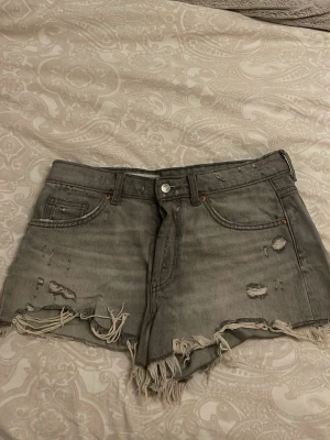 Grå slitna jeansshorts med fransar - Säljer ett par grå jeansshorts med slitna detaljer och fransig kant. Shortsen har klassisk femficksmodell, hög midja och råa avslut. Perfekta för dig som gillar en edgy och avslappnad stil. Materialet är kraftig bomull och färgen är tvättad grå.