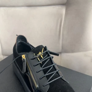 Svarta sneakers från Giuseppe Zanotti - Snygga svarta sneakers från Giuseppe Zanotti med lyxiga gulddetaljer, dragkedjor på sidorna och svart snörning. Skorna har en mix av mocka och lackdetaljer samt guldfärgad logga och häl. Perfekta för dig som vill sticka ut med exklusiv stil.