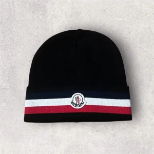 Moncler svart mössa med ränder - Snygg svart mössa från Moncler med breda ränder i marinblått, vitt och rött längs kanten. Mössan har Moncler-logga framtill och är stickad i mjukt material som håller dig varm. Perfekt för dig som vill ha en stilren accessoar med lyxig känsla.